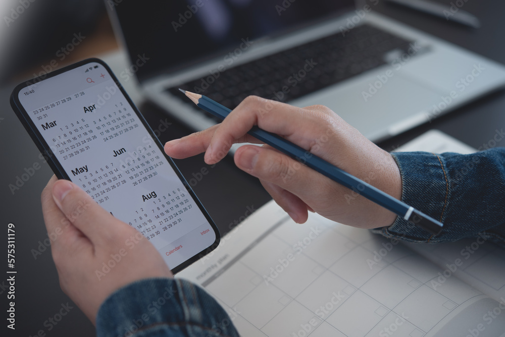 les mains d'une personne tenant dans la main droite un crayon papier et dans la main gauche un téléphone mobile connecté sur l'agenda google. Sous le bras posé sur la table un agenda papier, et à côté un PC